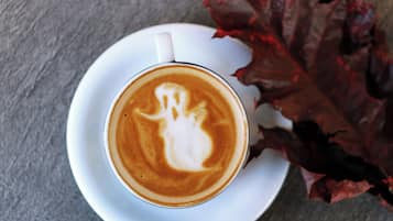 A cup of coffee with latte art on a white saucer, placed on a table with a red leaf beside it.