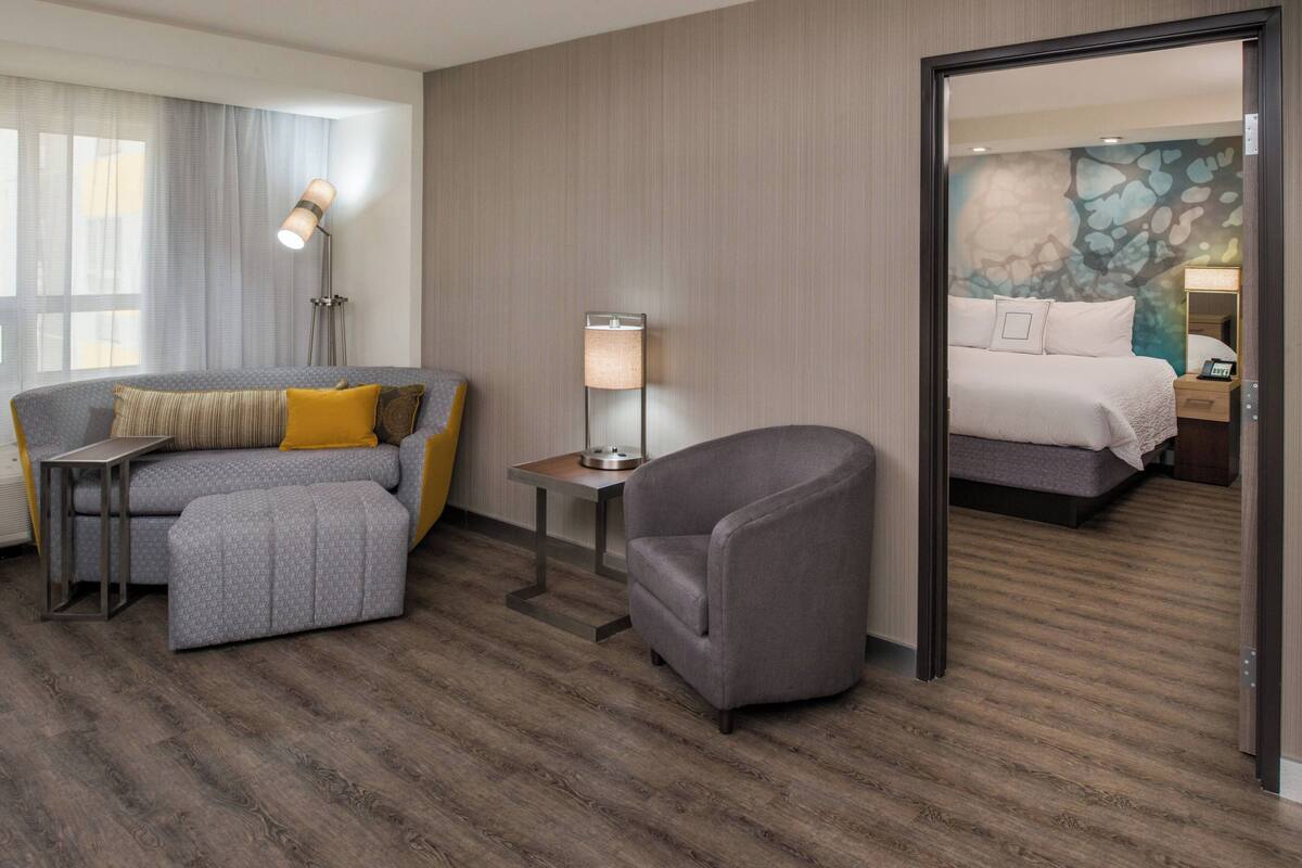 A hotel room with a grey sofa, a small armchair, a bedside table with a lamp, and a bed with white bedding.