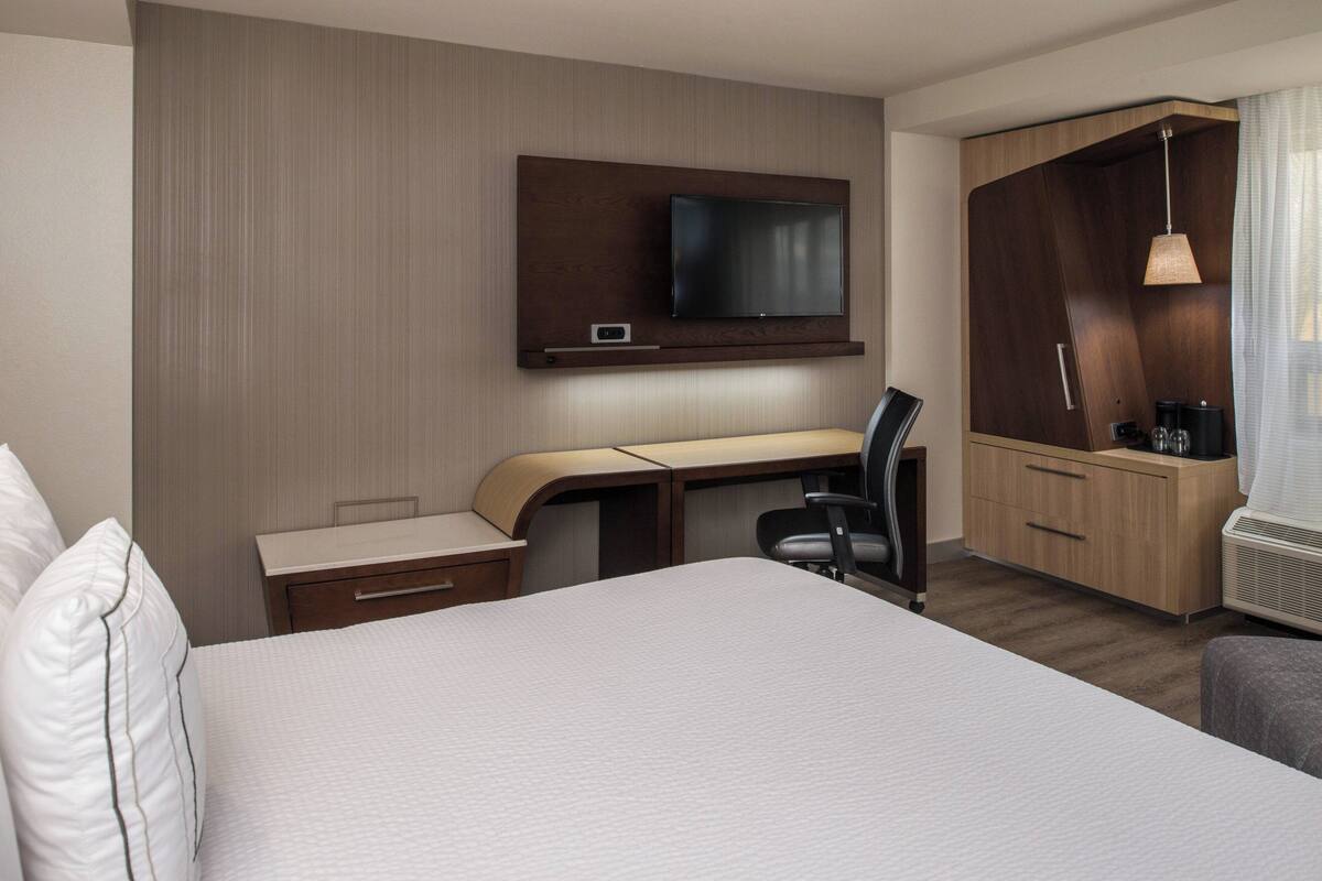 A hotel room with a bed, a desk with a chair, a television mounted on the wall, a wardrobe, and a small kitchenette area.