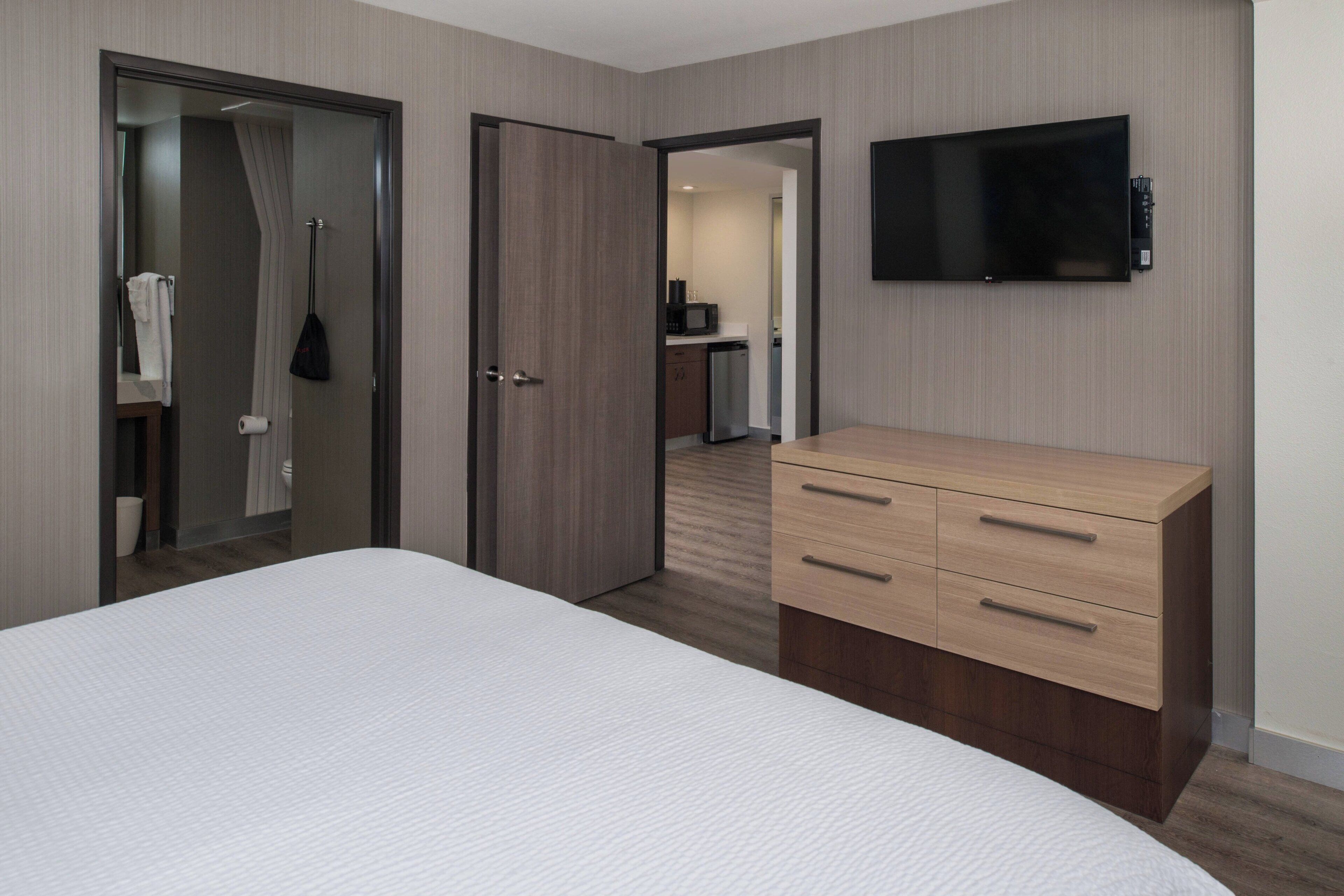 Een hotelkamer met een bed, een houten dressoir, een flatscreen televisie aan de muur en een deur naar een badkamer en een kitchenette.