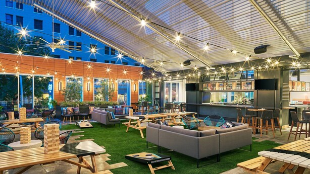 An outdoor seating area with wooden tables and benches, a gray sofa, and a bar area with a counter and stools. - Element by Marriott Austin Downtown (Austin)