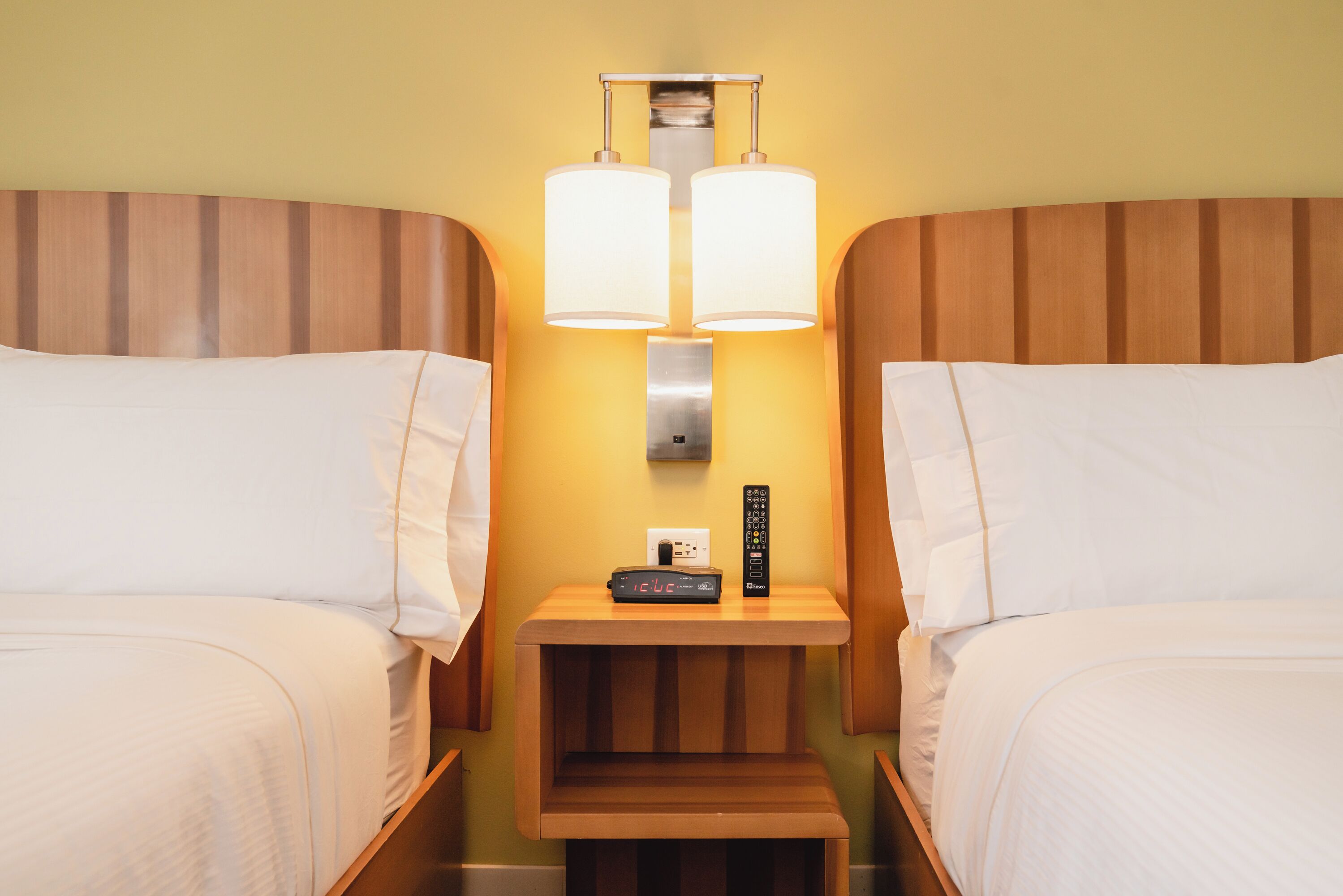 A hotel room with two beds, a nightstand with a clock and remote controls, and a wall-mounted lamp.