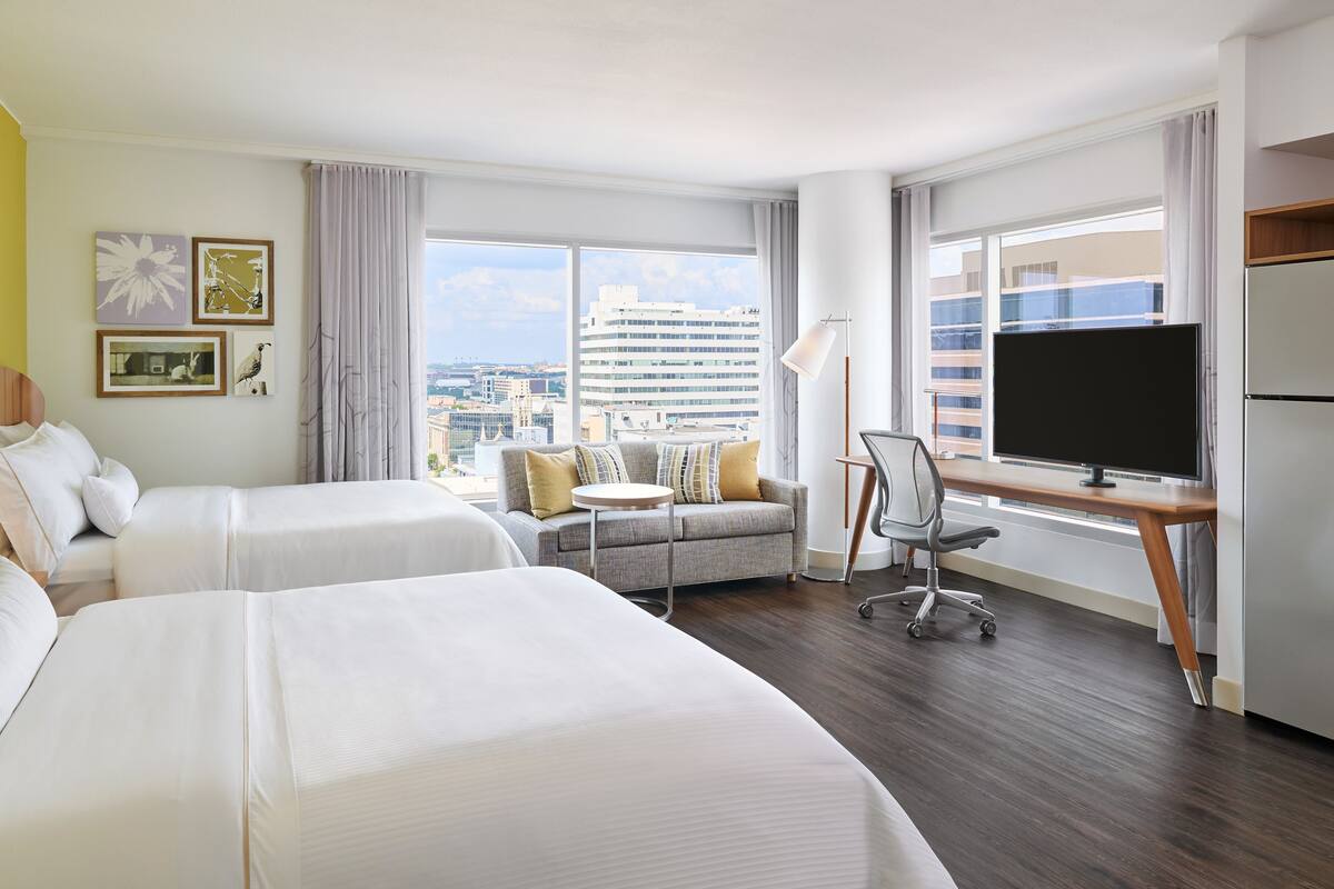 Habitación de hotel con dos camas, un escritorio con televisor, un sofá y vista a la ciudad.
