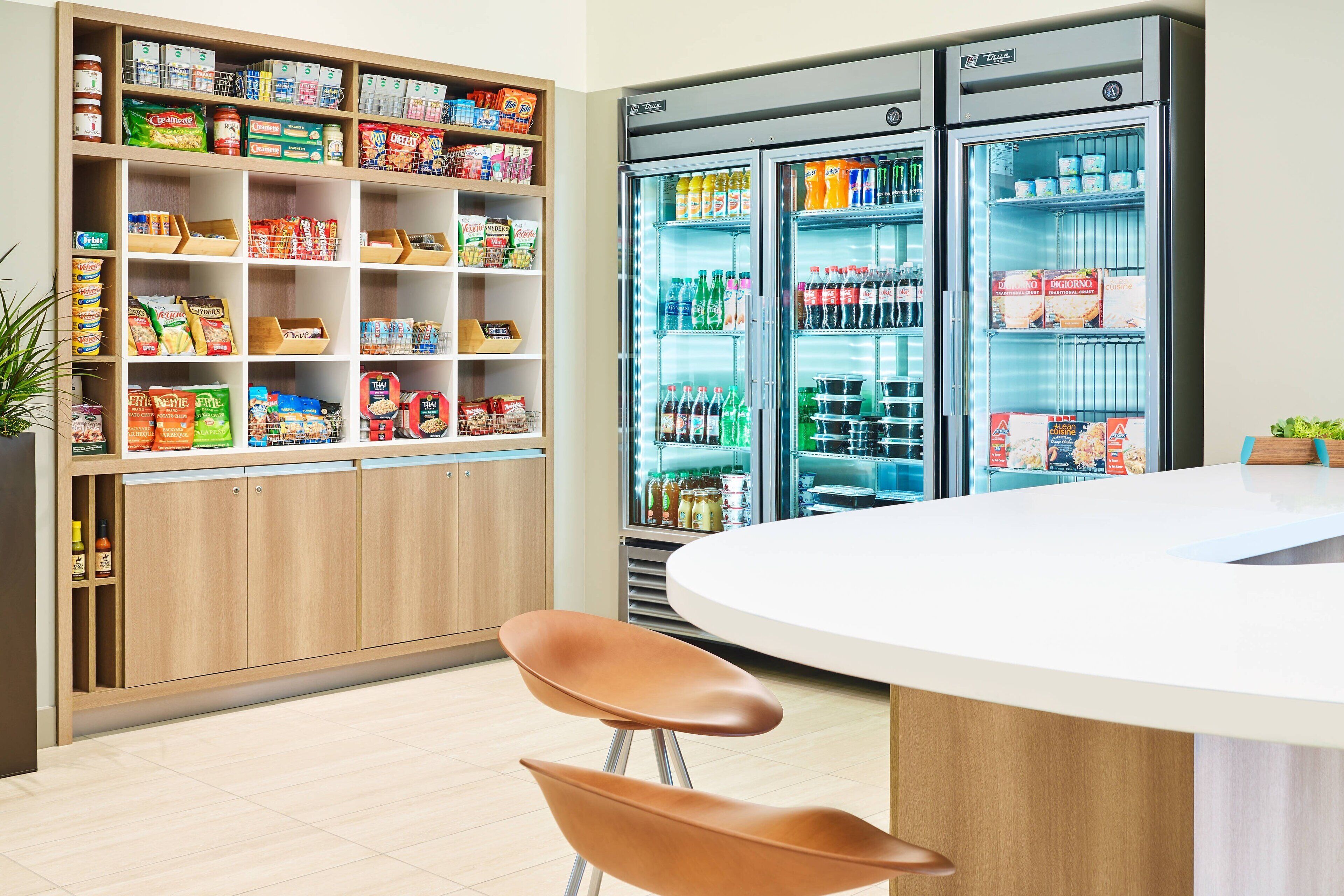 A modern kitchen with wooden cabinets, a white countertop, and two brown chairs. There are two refrigerators stocked with beverages and food items.