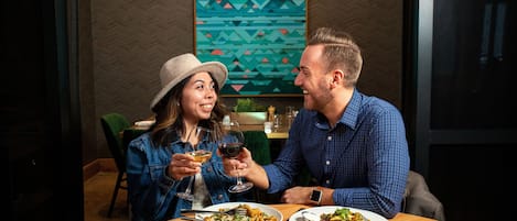 Un couple dînant dans un restaurant décoré d’œuvres d’art géométriques murales, avec une table en bois et un éclairage suspendu.