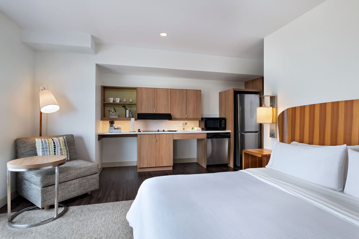 Una habitación de hotel pequeña con kitchenette, una cama, un rincón de descanso y una lámpara.