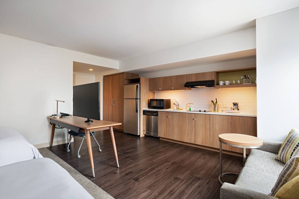 Una cocina moderna con armarios de madera, un microondas empotrado y un refrigerador de acero inoxidable. Hay una mesa de comedor y una mesita redonda con una silla.