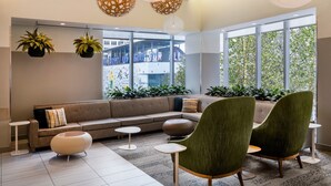 A modern lounge area with a sofa, two green armchairs, a round white ottoman, and hanging planters. - Element by Marriott Austin Downtown (Austin)