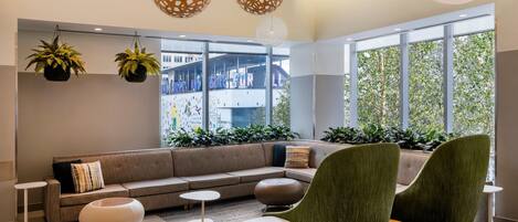 A modern lounge area with a sofa, two green armchairs, a round white ottoman, and hanging planters.
