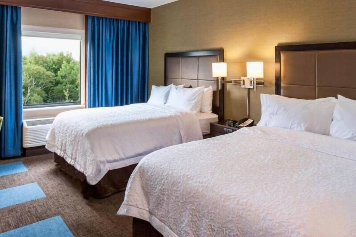 A hotel room with two beds, a nightstand with a phone, a window with curtains, and a view of greenery outside.