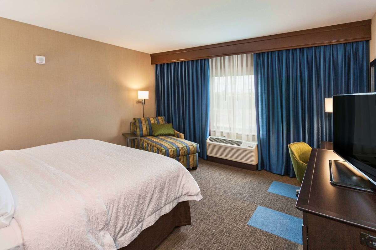 A hotel room with a large bed, a desk with a TV, a chair, a lamp, and a window with curtains.