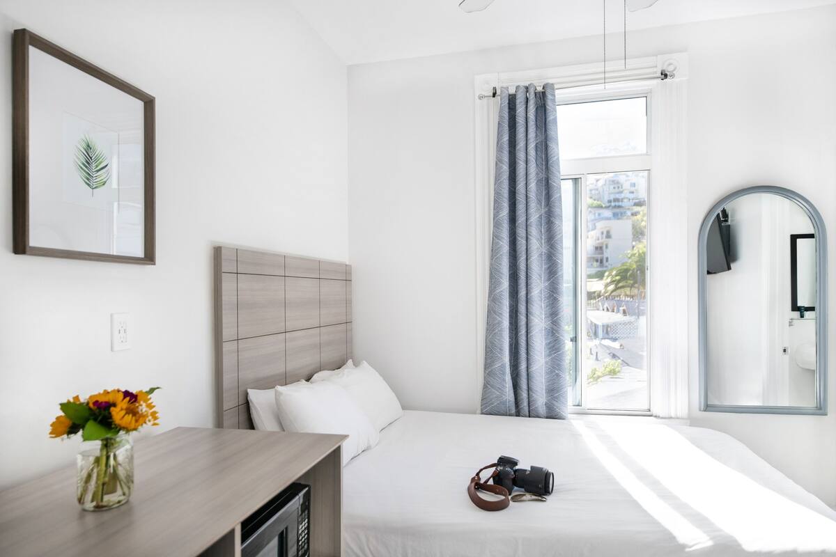 Habitación de hotel con una cama, una mesita de noche con un florero, una cámara y una ventana con cortinas.