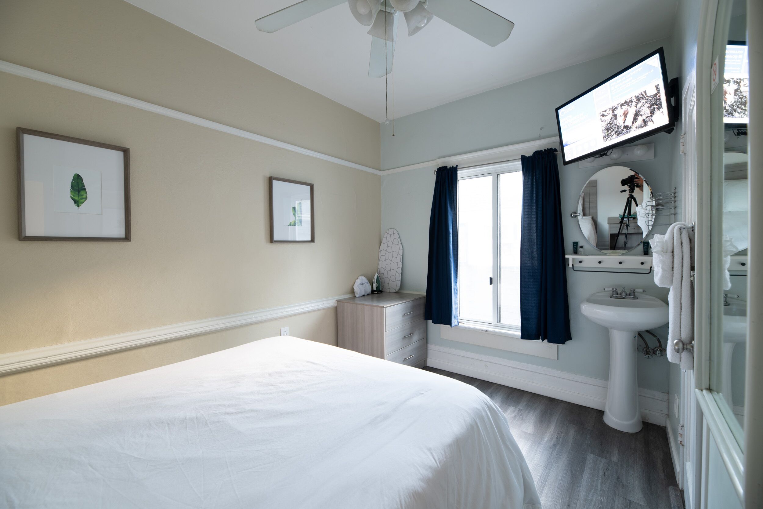 A bedroom with a bed, a dresser, a window with curtains, a television, and a bathroom with a sink and mirror.