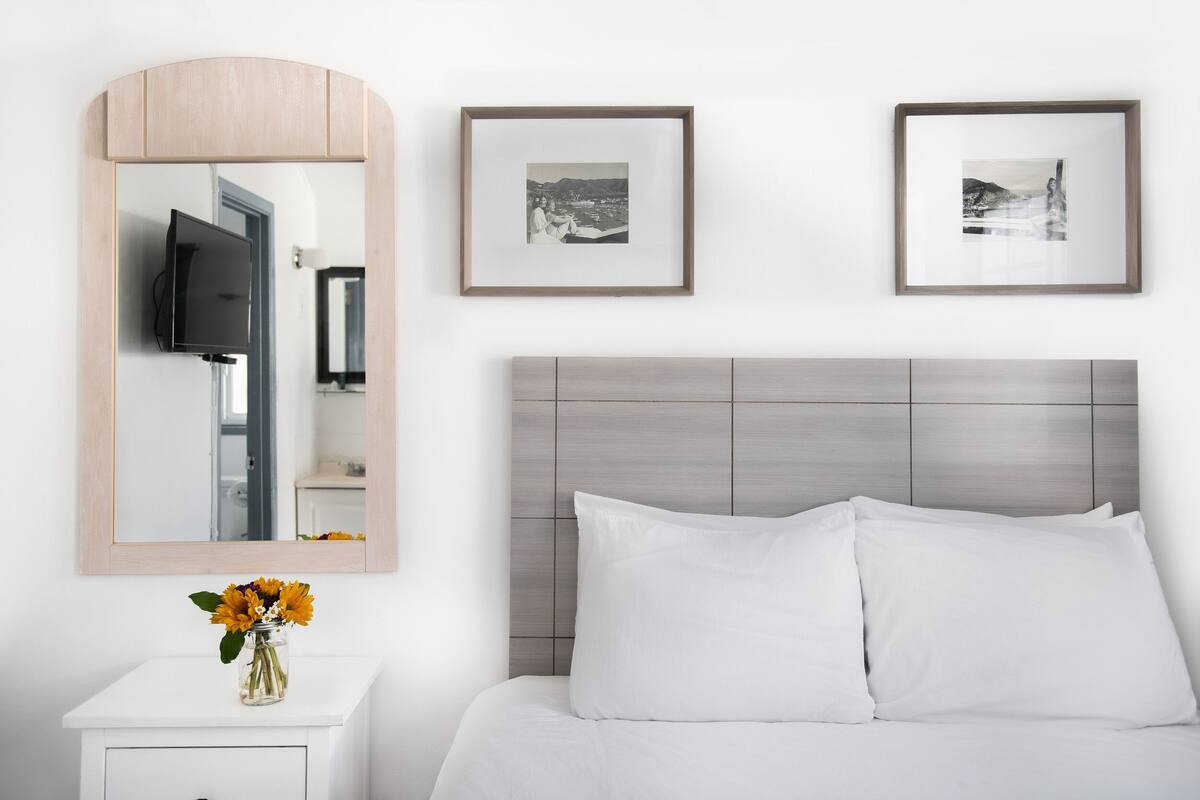 Habitación de hotel con cama, televisor sobre un pedestal, espejo y dos cuadros en la pared.