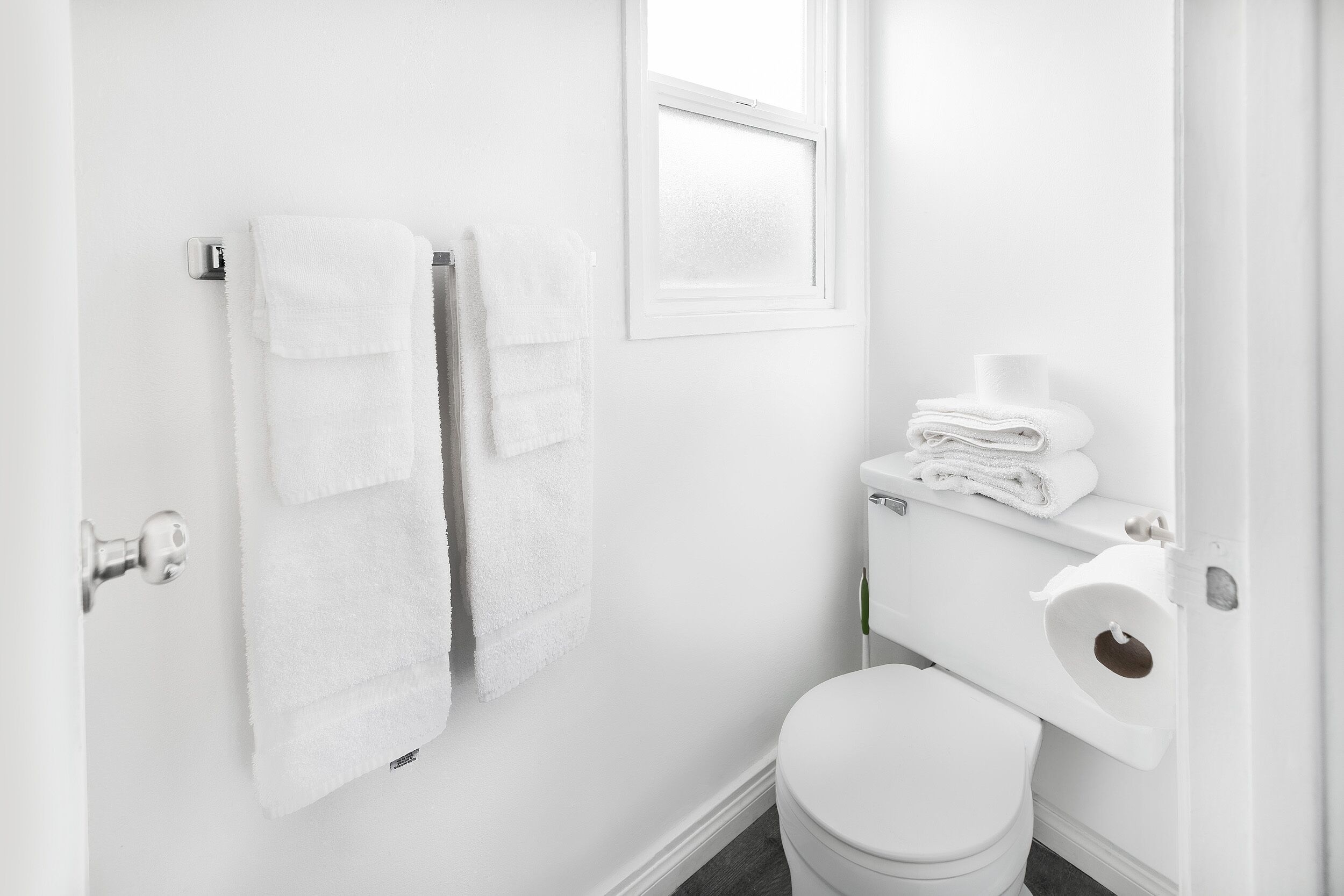 A white bathroom with a toilet, a towel rack with folded towels, and a window.