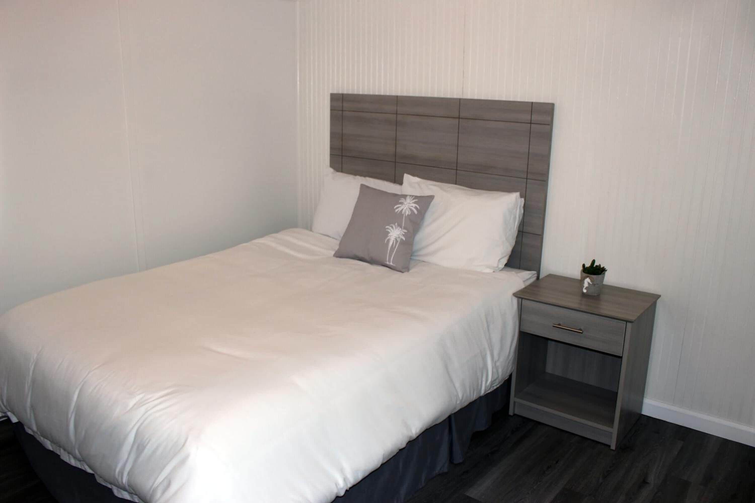 A neatly made bed with a grey headboard, a bedside table with a plant, and a wall with vertical lines.