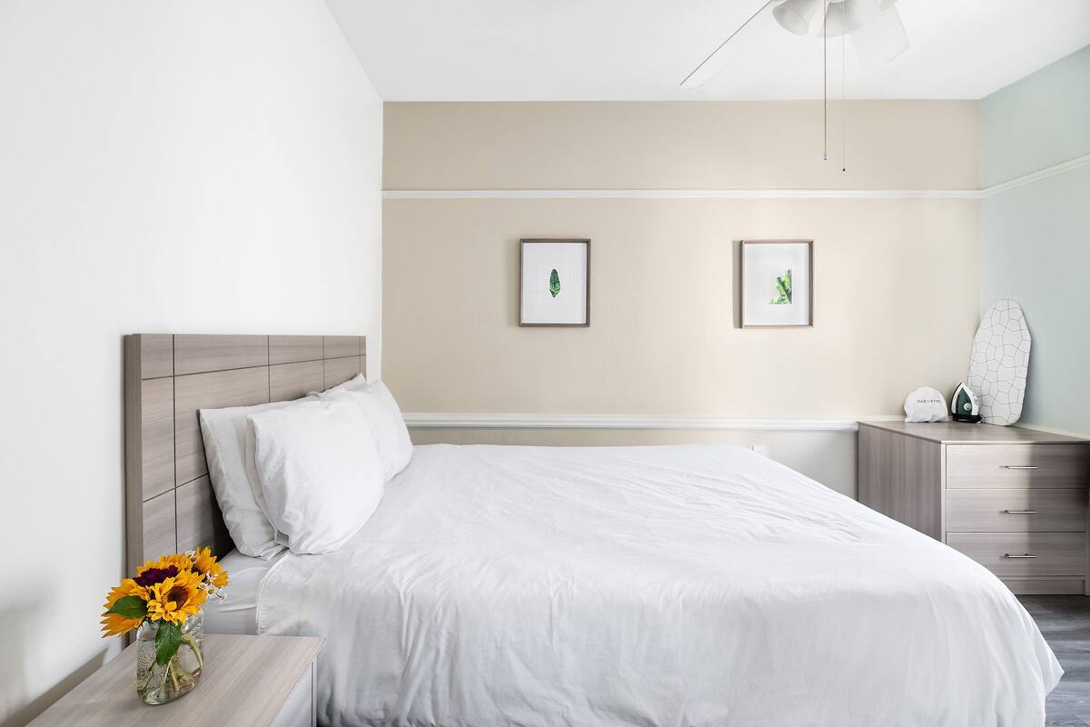 Habitación de hotel impecable con una cama, una mesita de noche con un jarrón de girasoles y dos cuadros en la pared.