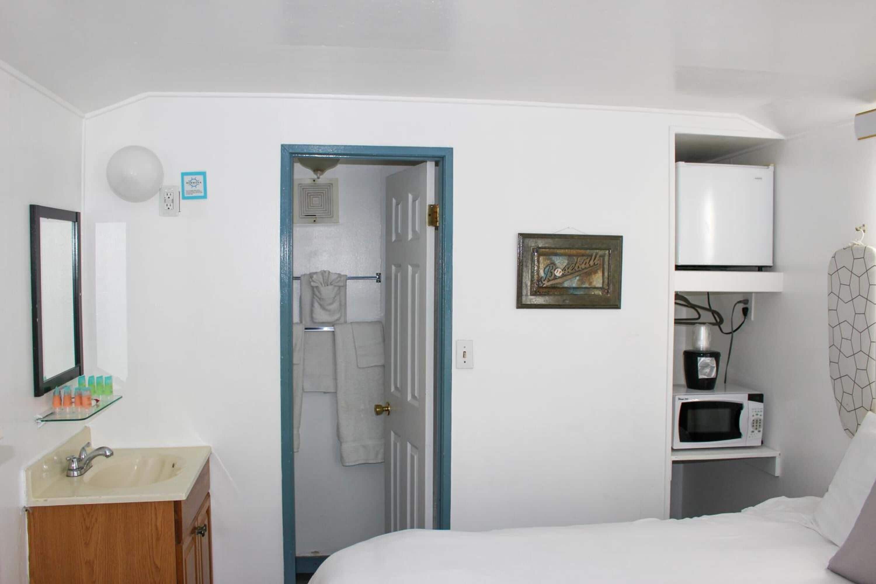 A bedroom with a bed, a white wardrobe, a bathroom with a sink and mirror, and a door leading to another room.