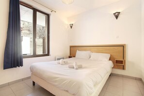 A hotel room with a large bed, a window with curtains, a bedside table, and a wall-mounted light fixture. - Cozy Balcony Apartment (La Clusaz)