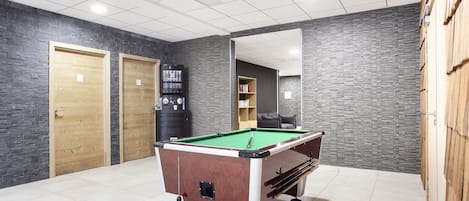 Une table de billard dans une pièce dont le sol est carrelé, qui possède des portes en bois et un mur à la texture particulière.