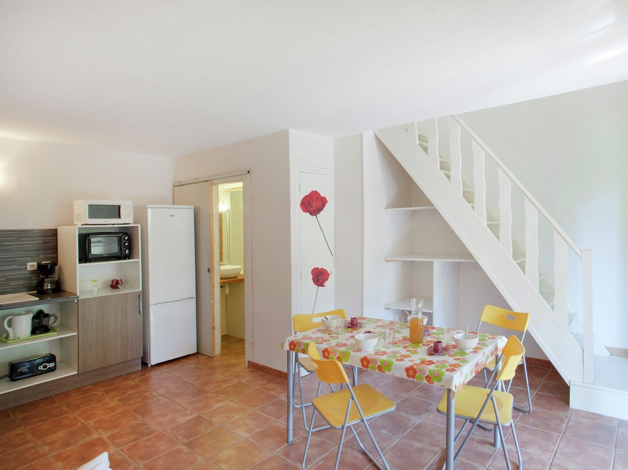 Eine kompakte Küche mit Mikrowelle, Toaster und Kühlschrank. Ein Esstisch mit gelben Stühlen und einer Tischdecke mit Blumenmuster befindet sich in der Mitte. Eine weiße Treppe ist rechts zu sehen.