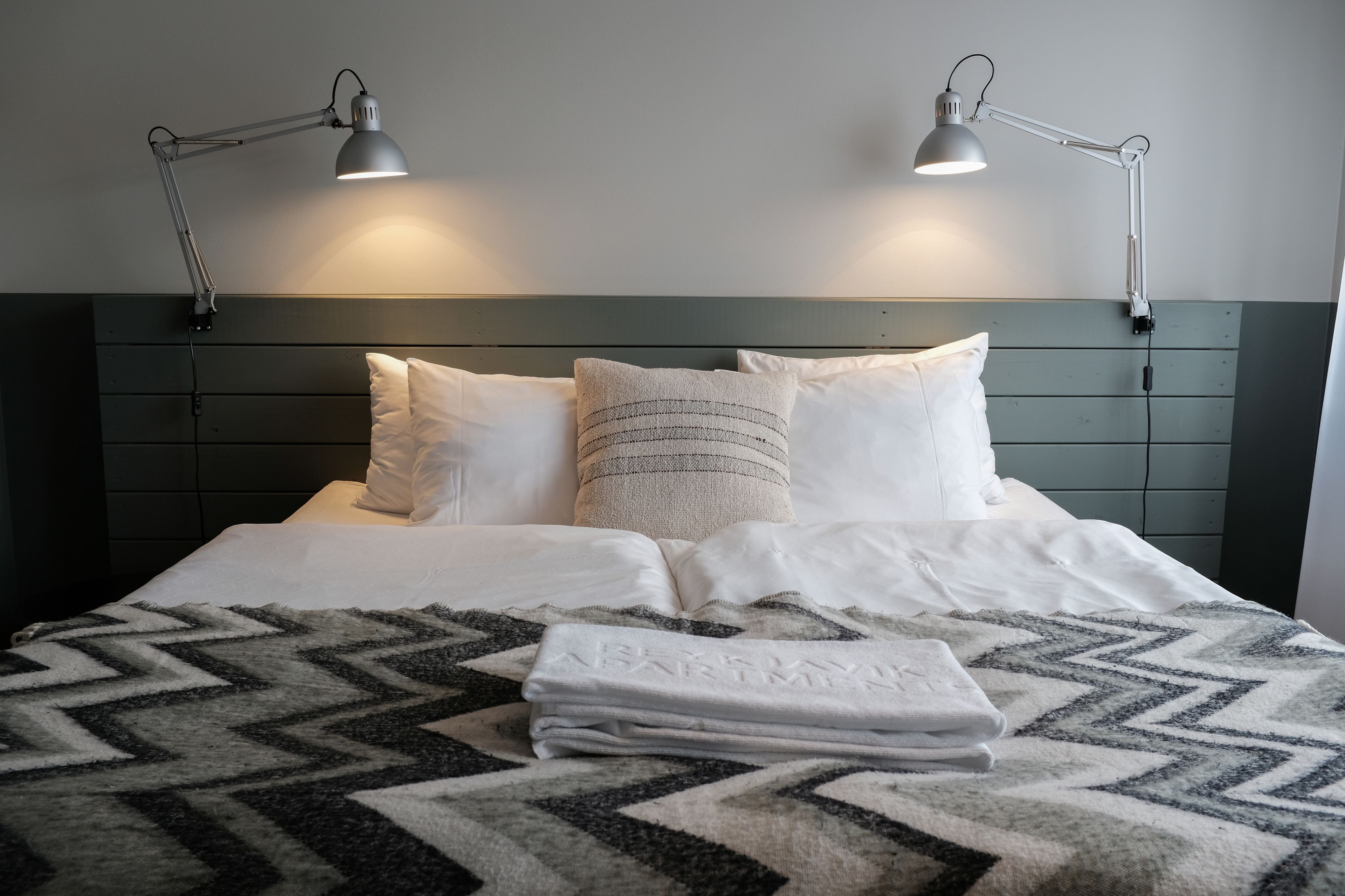 A neatly made bed with two pillows, a folded towel, and two bedside lamps on a wall-mounted headboard.