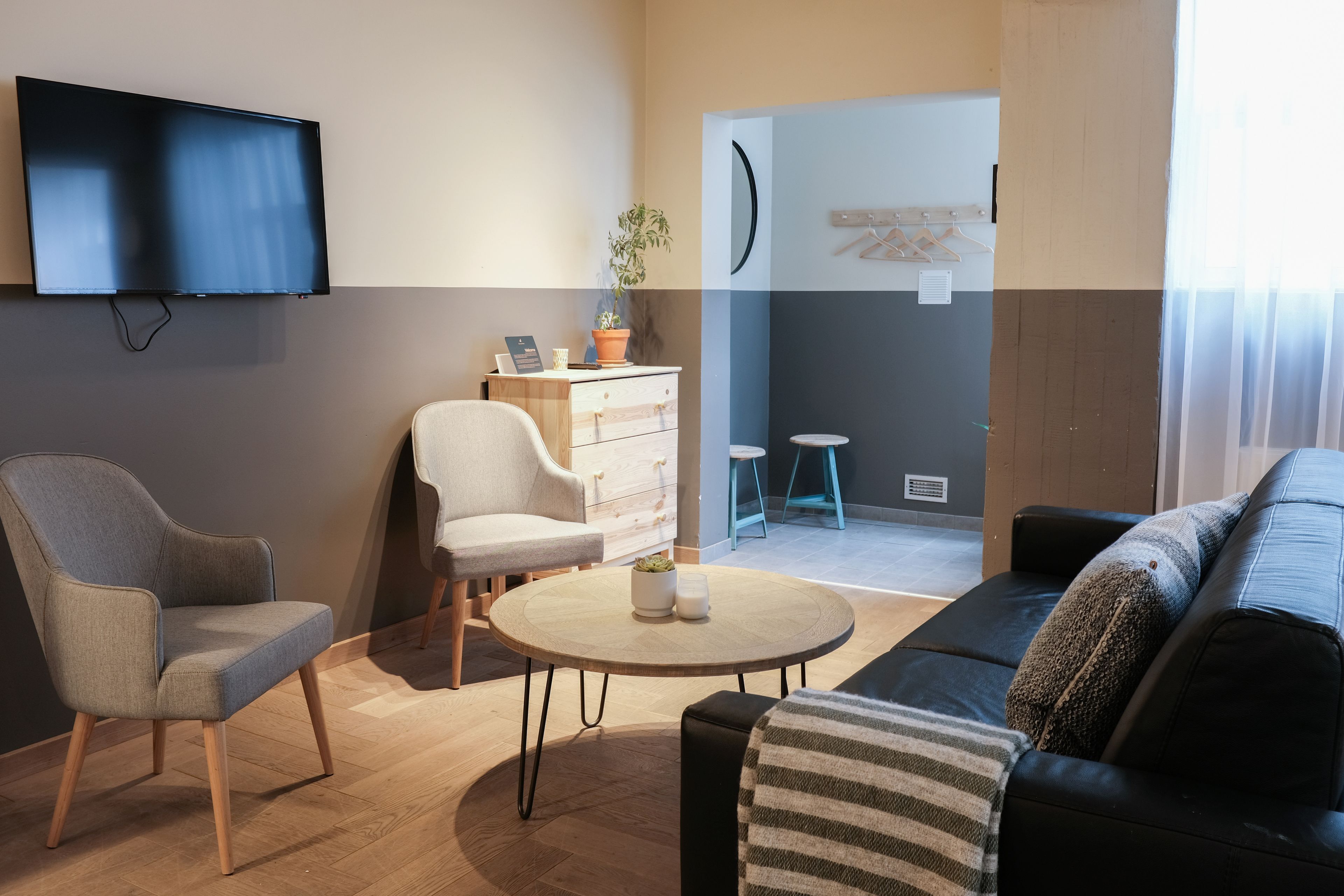 Un saló modern amb televisió de pantalla plana, un butac armchair gris, una cadira blanca, una taula de centre rodona de fusta i un sofà negre amb coixins a ratlles.