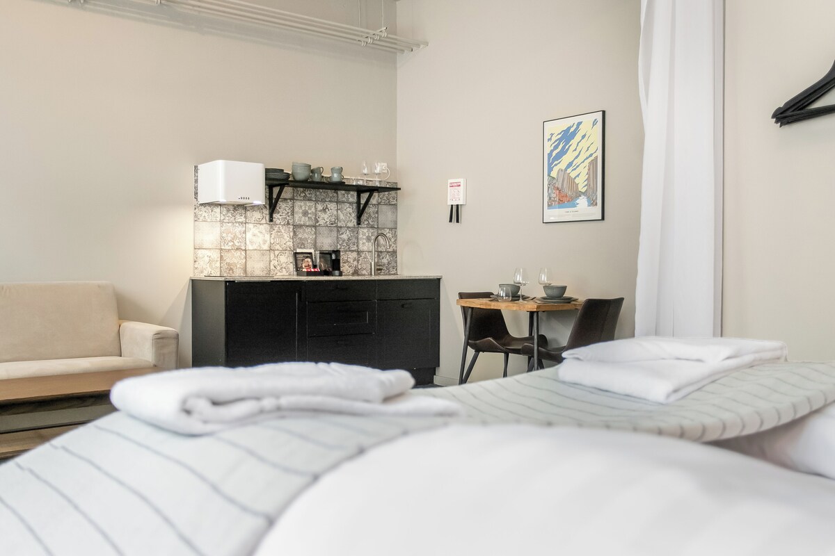 A hotel room with a bed, a small desk with a chair, a kitchenette, and a framed picture on the wall.