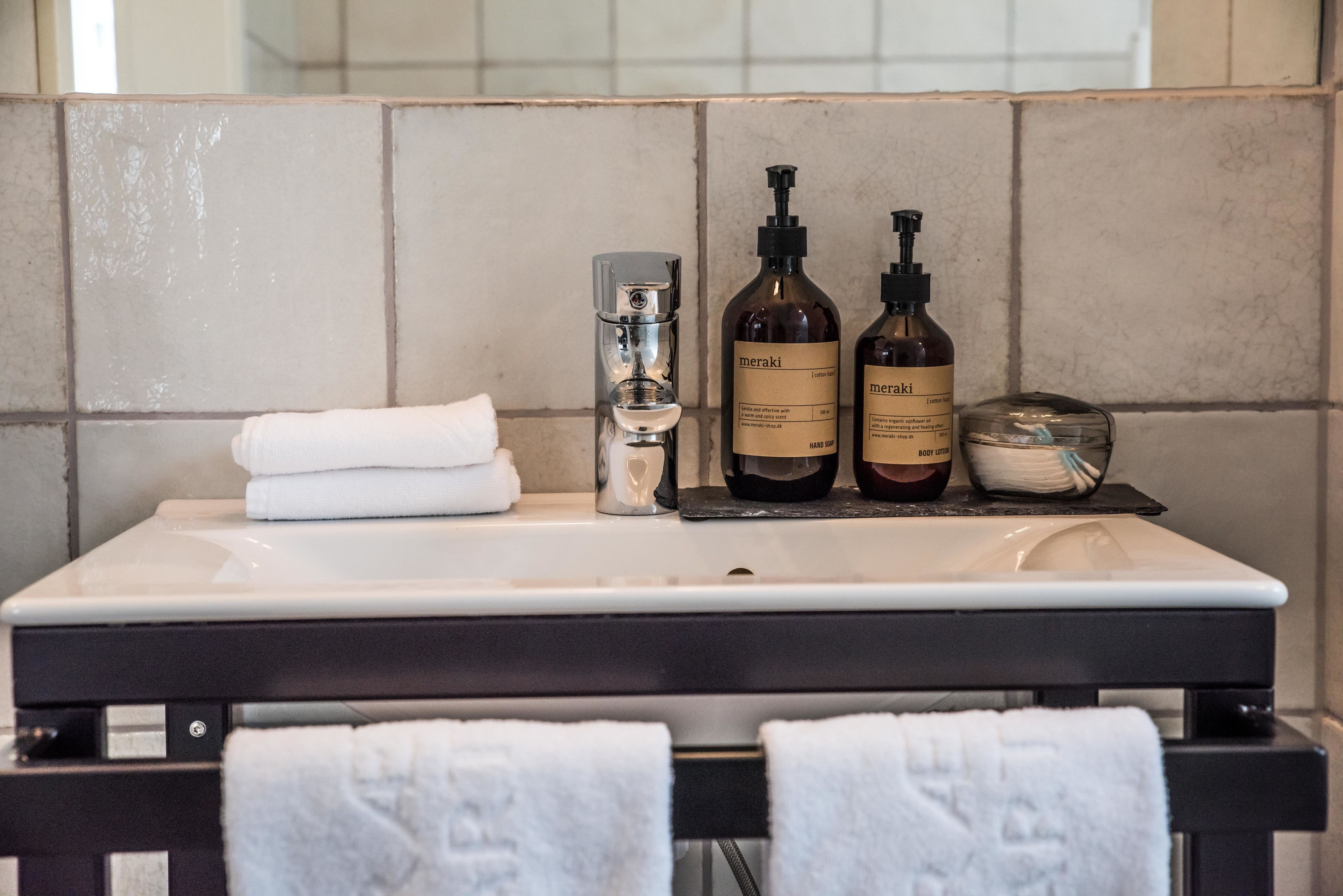 A bathroom with a white sink, two Meraki body lotion bottles, a glass soap dispenser, and a white towel on the counter.
