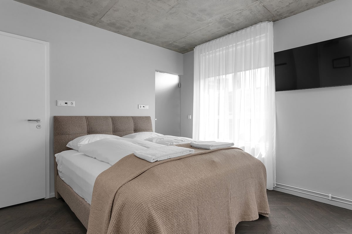 A modern bedroom with a bed, a flat-screen TV, and a window with sheer curtains.