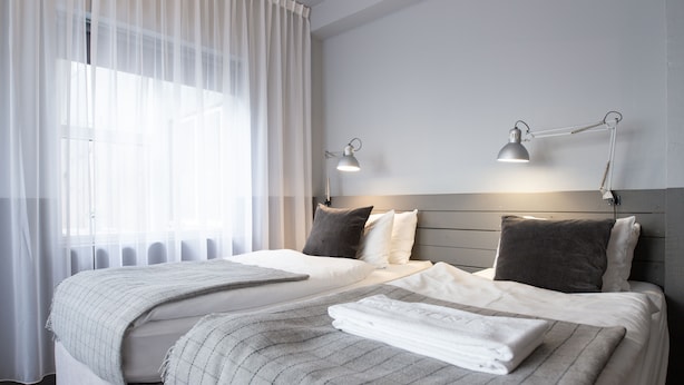 A neatly made double bed with a grey checkered blanket and pillows, two bedside lamps, and a window with sheer curtains.