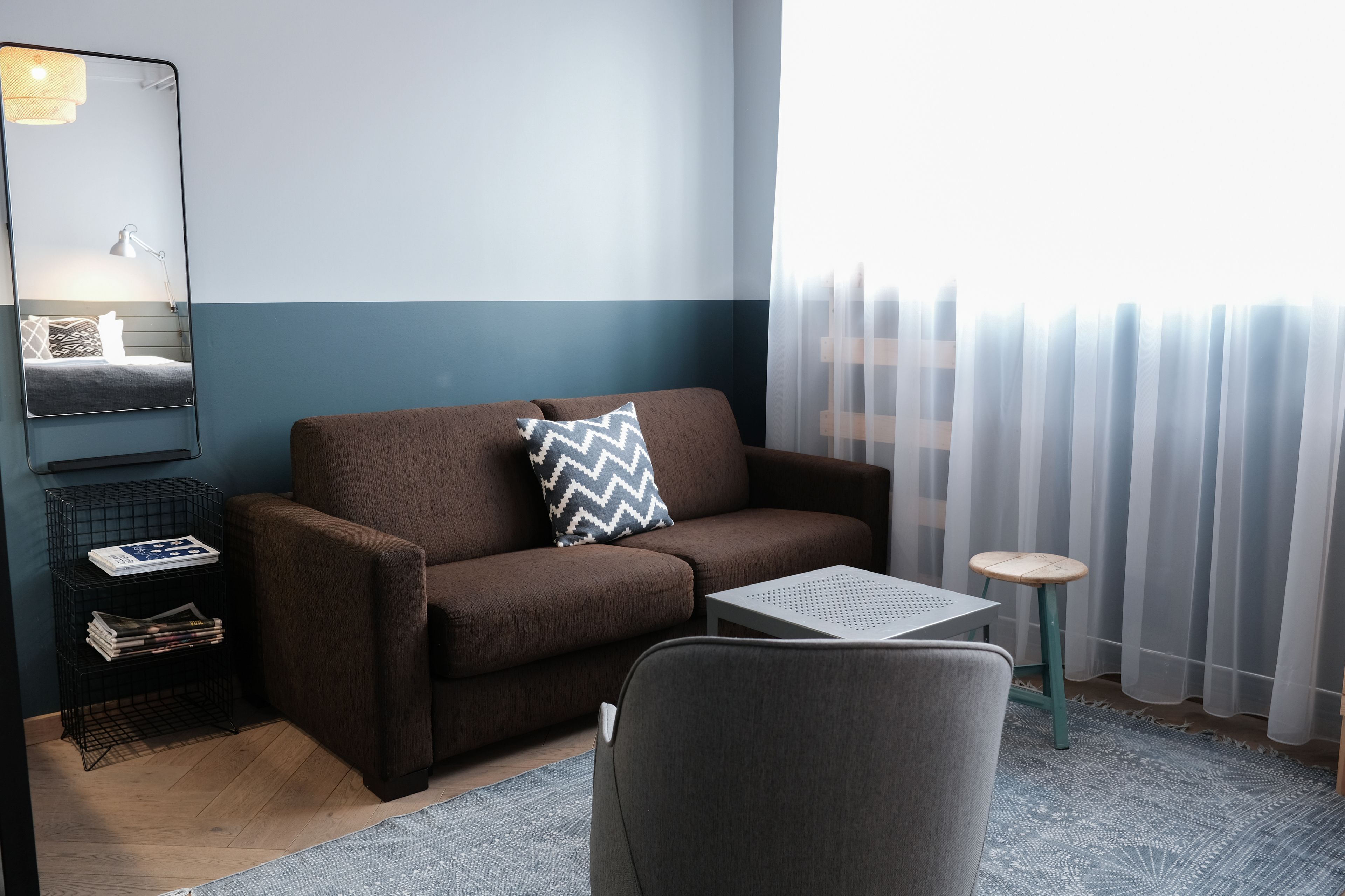 A modern living room with a brown sofa, a small table, a chair, a side table with magazines, and a mirror on the wall.