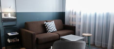A modern living room with a brown sofa, a small table, a chair, a side table with magazines, and a mirror on the wall.