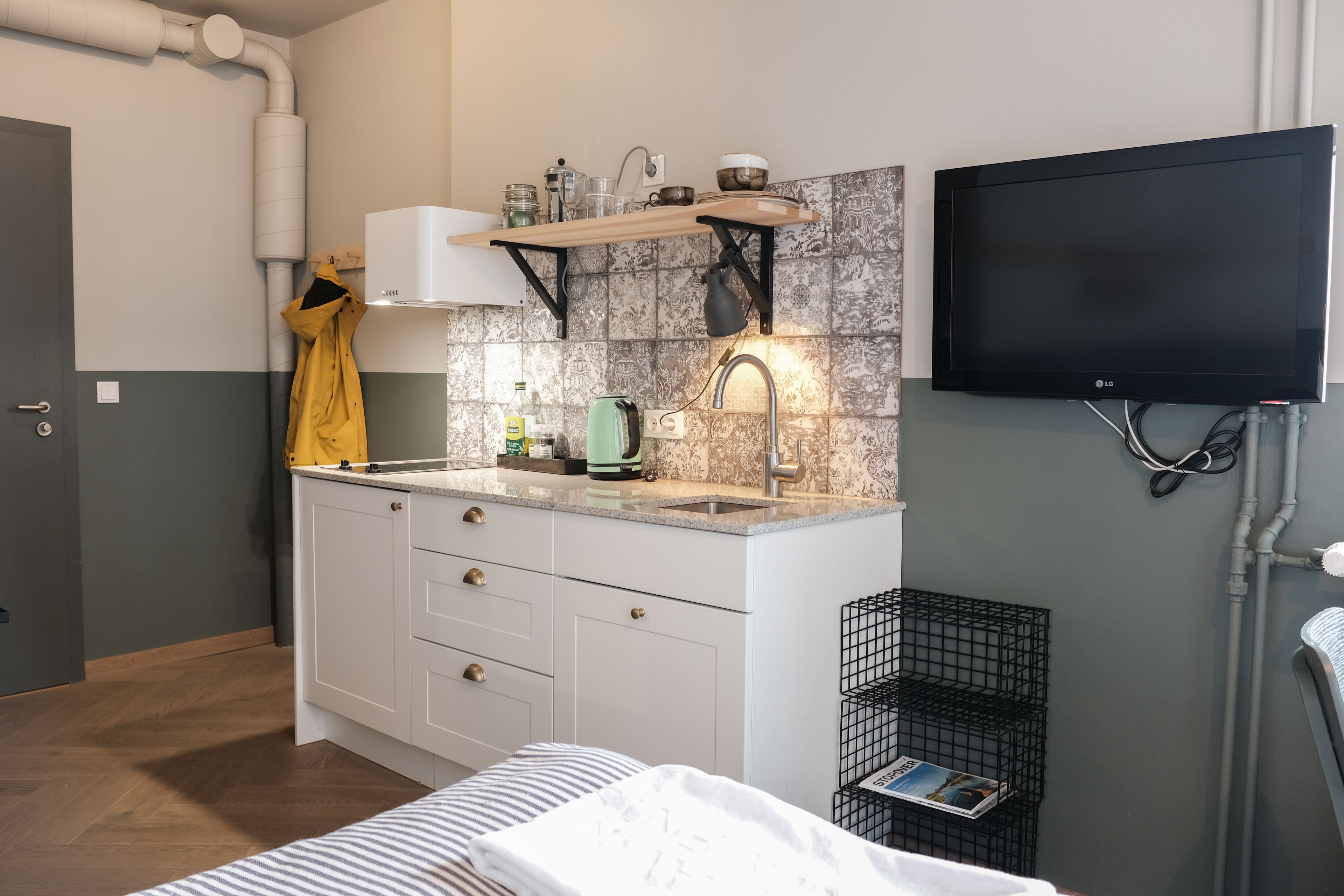 Cocina compacta con estufa integrada, lavamanos y una pequeña encimera. Hay un televisor montado en la pared, un estante sobre el lavamanos y un soporte con utensilios de cocina.