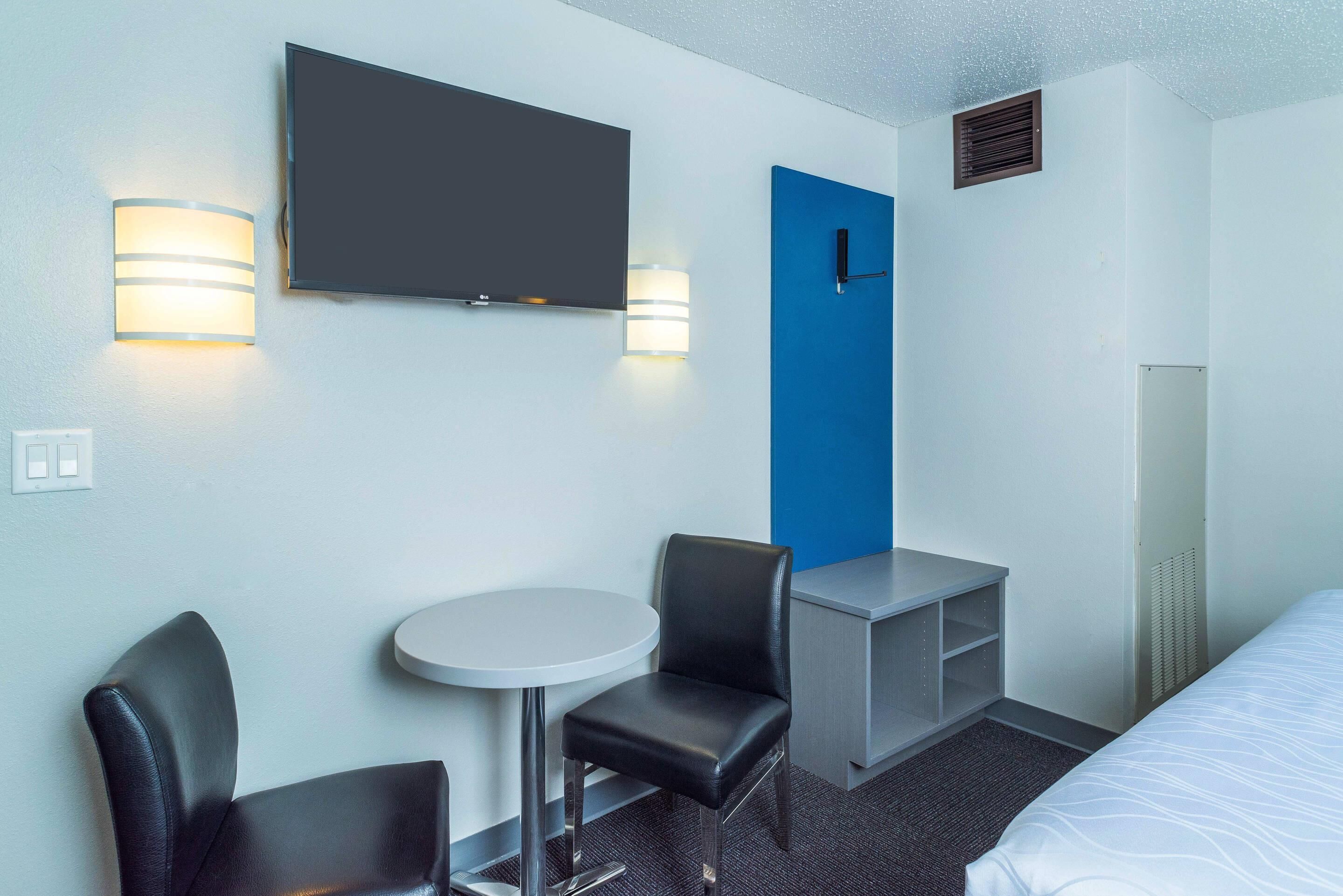A hotel room with a bed, a desk, two chairs, a TV, and wall-mounted lights.