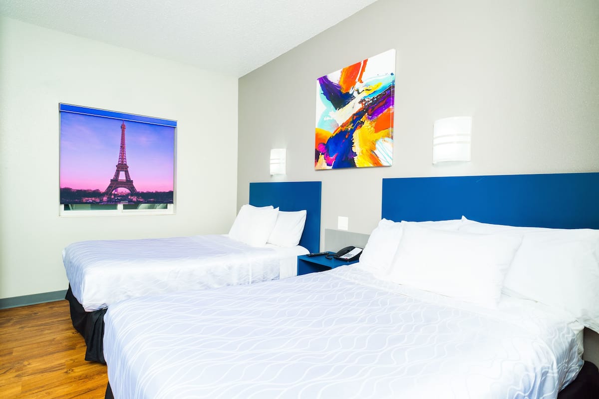 A hotel room with two beds, a blue headboard, a painting of the Eiffel Tower, and a colorful abstract painting.