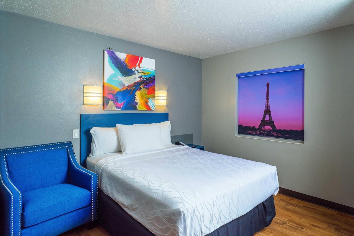 A hotel room with a bed, a blue armchair, a colorful abstract painting, a picture of the Eiffel Tower, and a wall-mounted lamp.