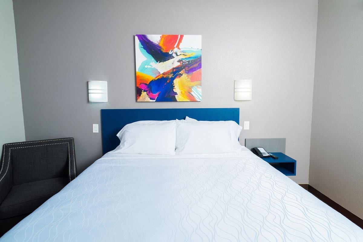 A hotel room with a bed, a chair, a nightstand, and a colorful abstract painting on the wall.