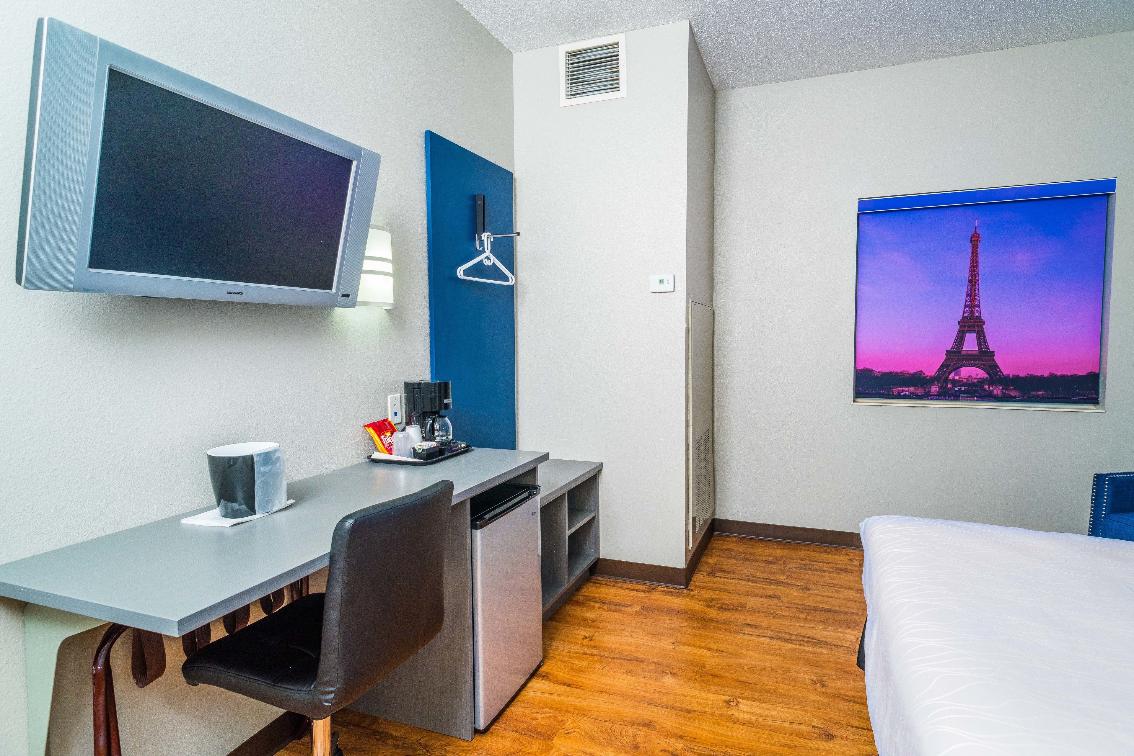 A hotel room with a bed, a desk with a computer, a small fridge, a TV, and a picture of the Eiffel Tower.
