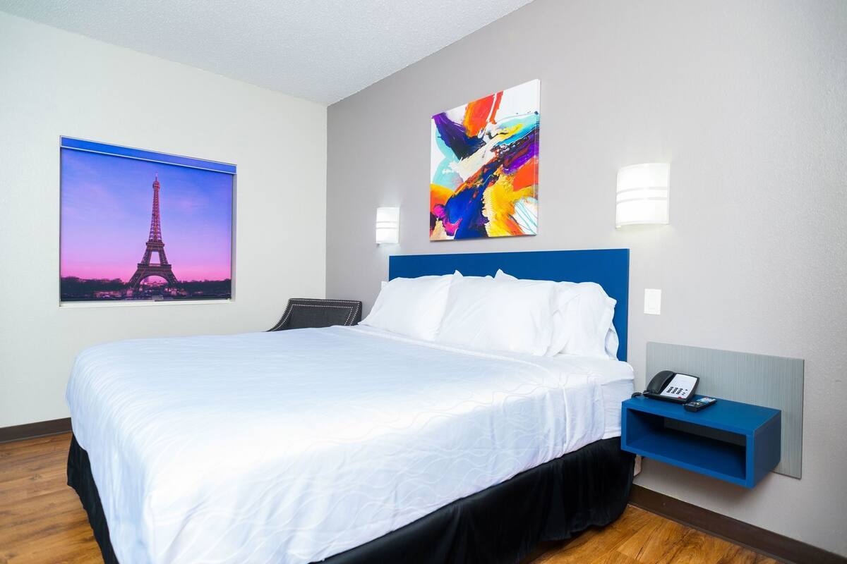 A hotel room with a bed, a nightstand with a phone, a wall-mounted light, and two paintings.