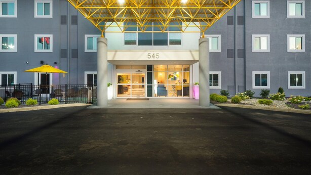 Entrada moderna de un hotel con un toldo de vidrio geométrico, número 545, y un área exterior bien iluminada con asientos y plantas.