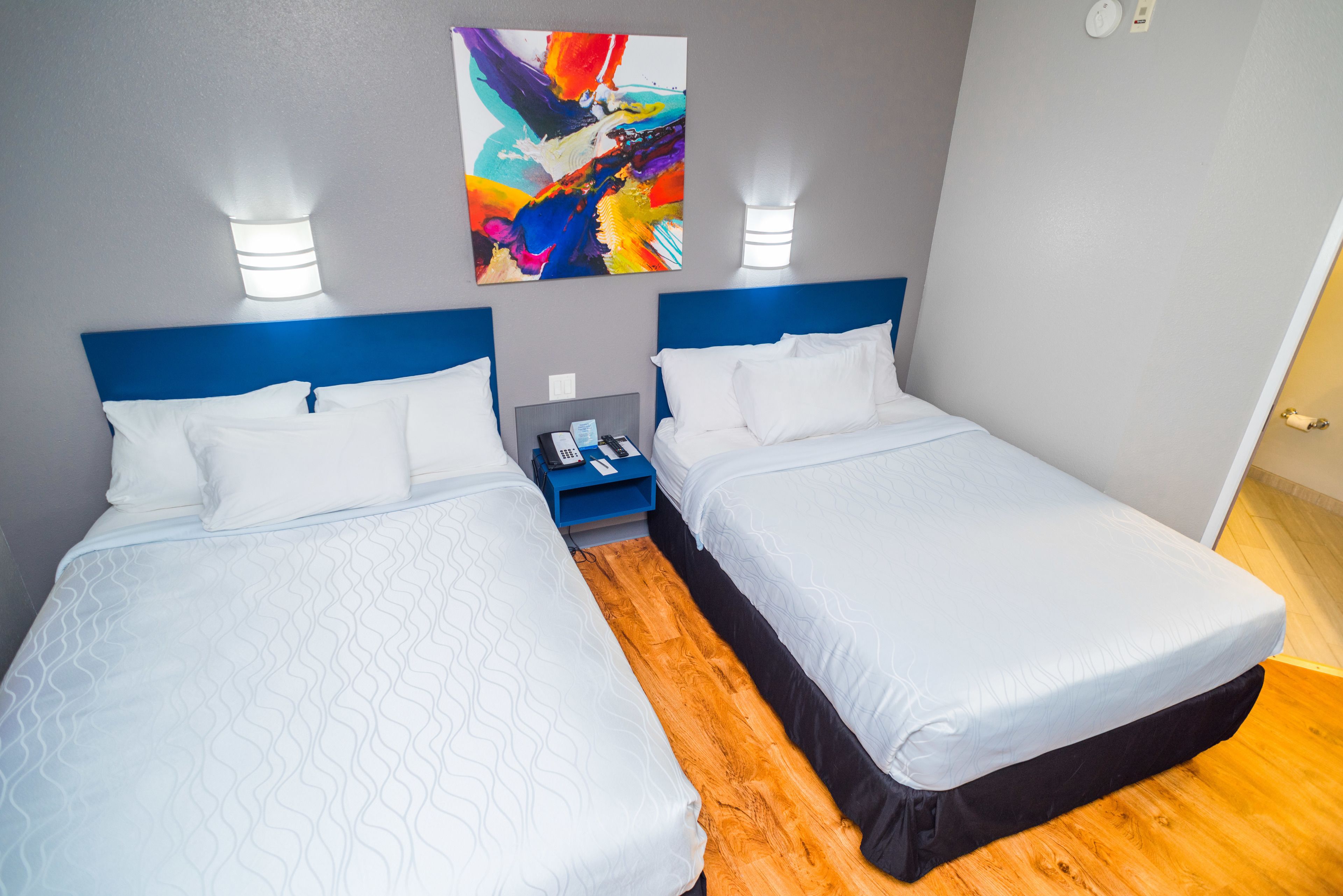 A hotel room with two beds, a blue headboard, white bedding, a colorful abstract painting on the wall, and a bedside table with a phone and a lamp.