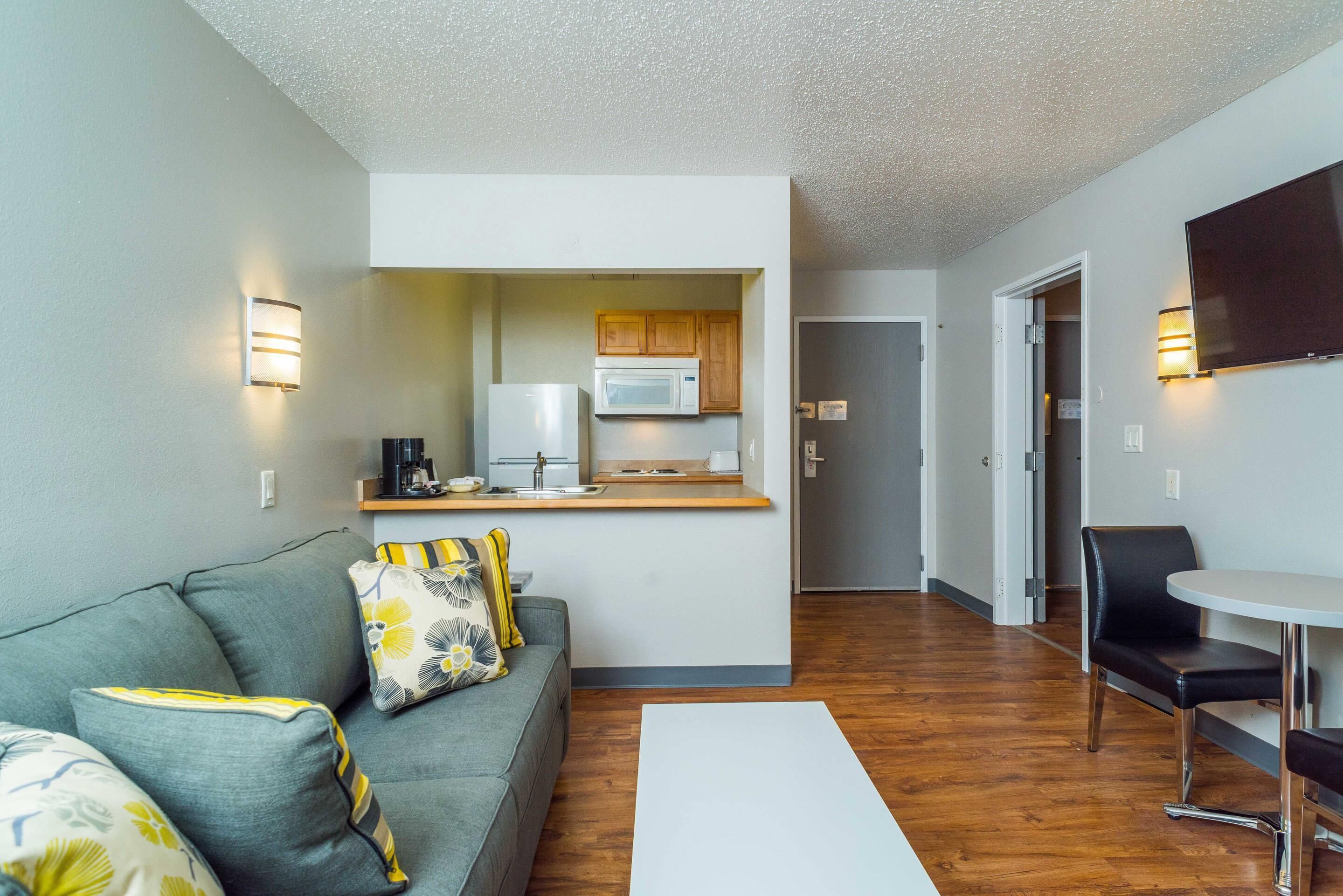 A compact living space with a kitchenette, a grey sofa with floral cushions, a small dining table, and a flat-screen TV mounted on the wall.