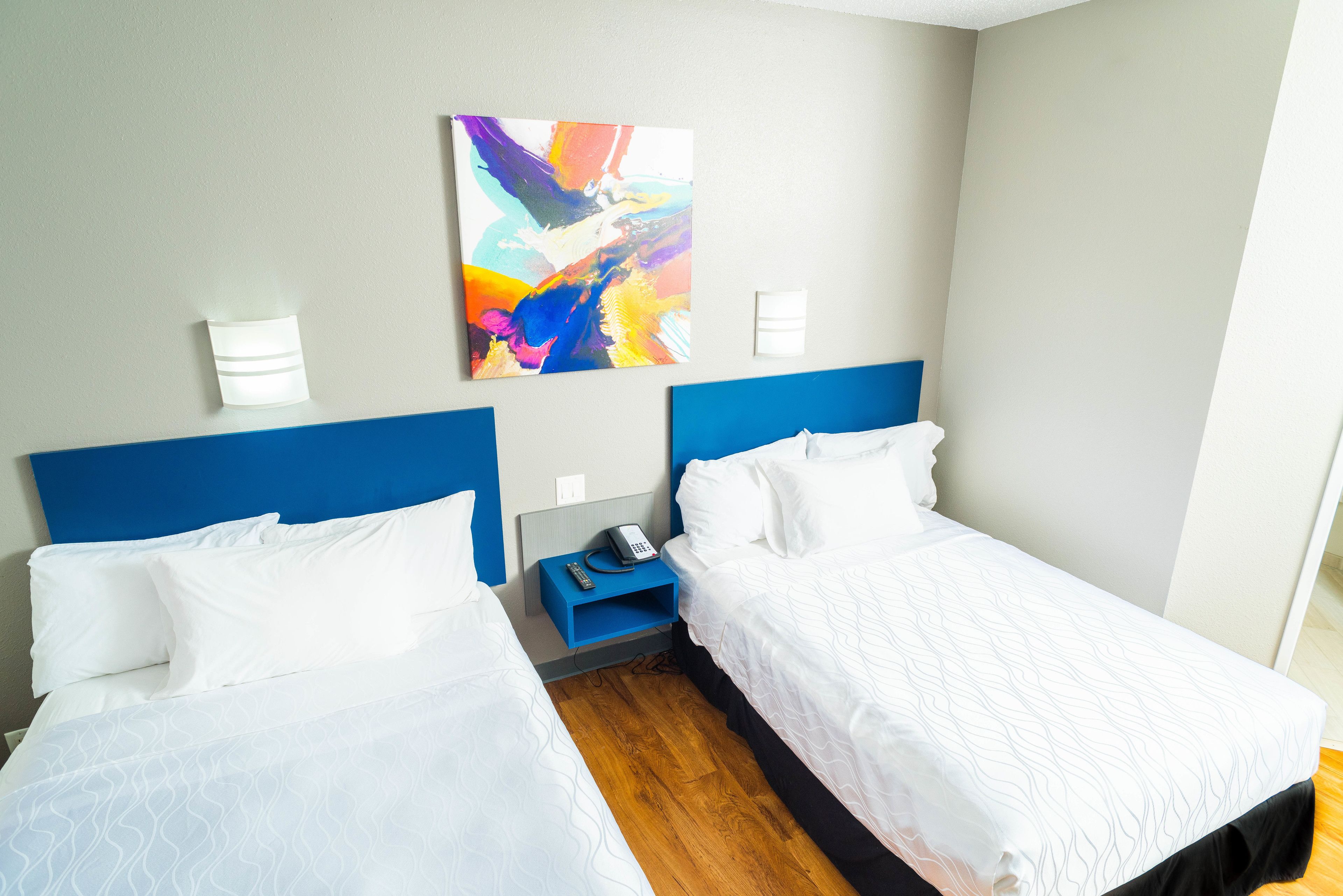 Habitación de hotel con dos camas, un cabecero azul, un cuadro abstracto de colores, una mesita de noche con teléfono y un estante azul pequeño.