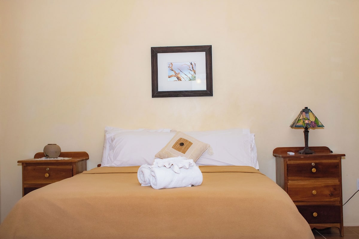 A neatly made bed with a brown bedspread, a rolled white towel, and a beige pillow. There is a framed picture on the wall above the bed and a small table with a lamp beside the bed.