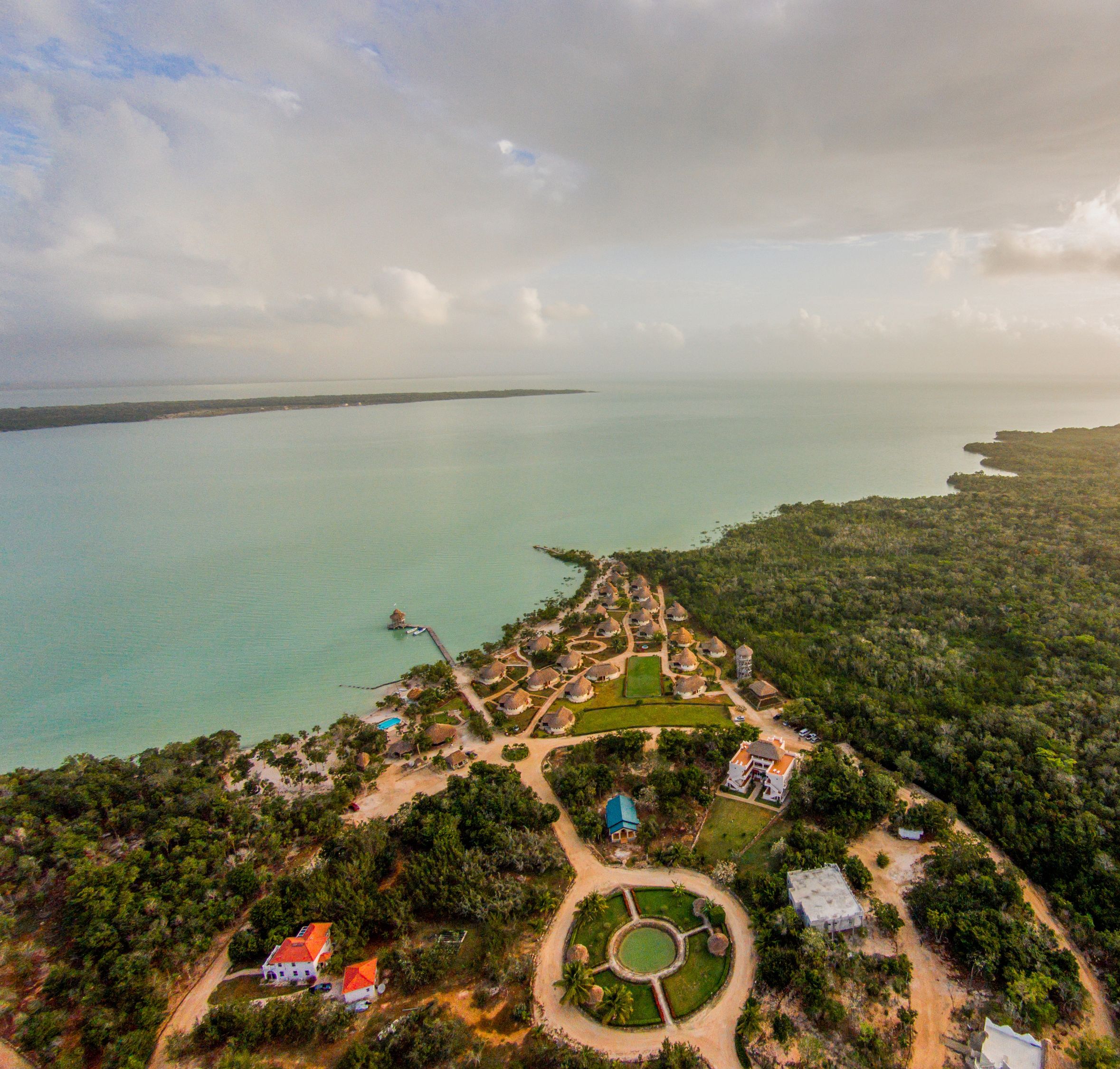 An aerial view of a coastal resort with a central circular garden, multiple bungalow-style buildings, and a pier extending into the sea.
