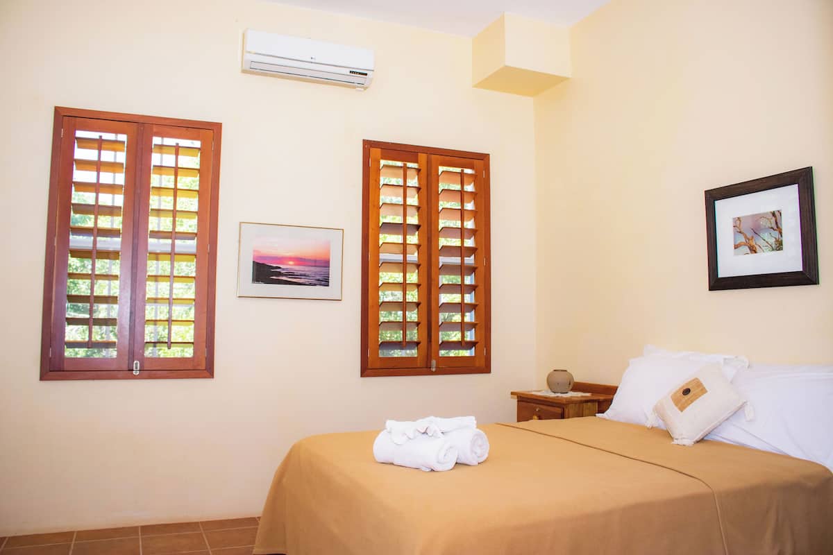A hotel room with a bed, two windows with wooden shutters, a wall-mounted air conditioner, and a framed picture on the wall.