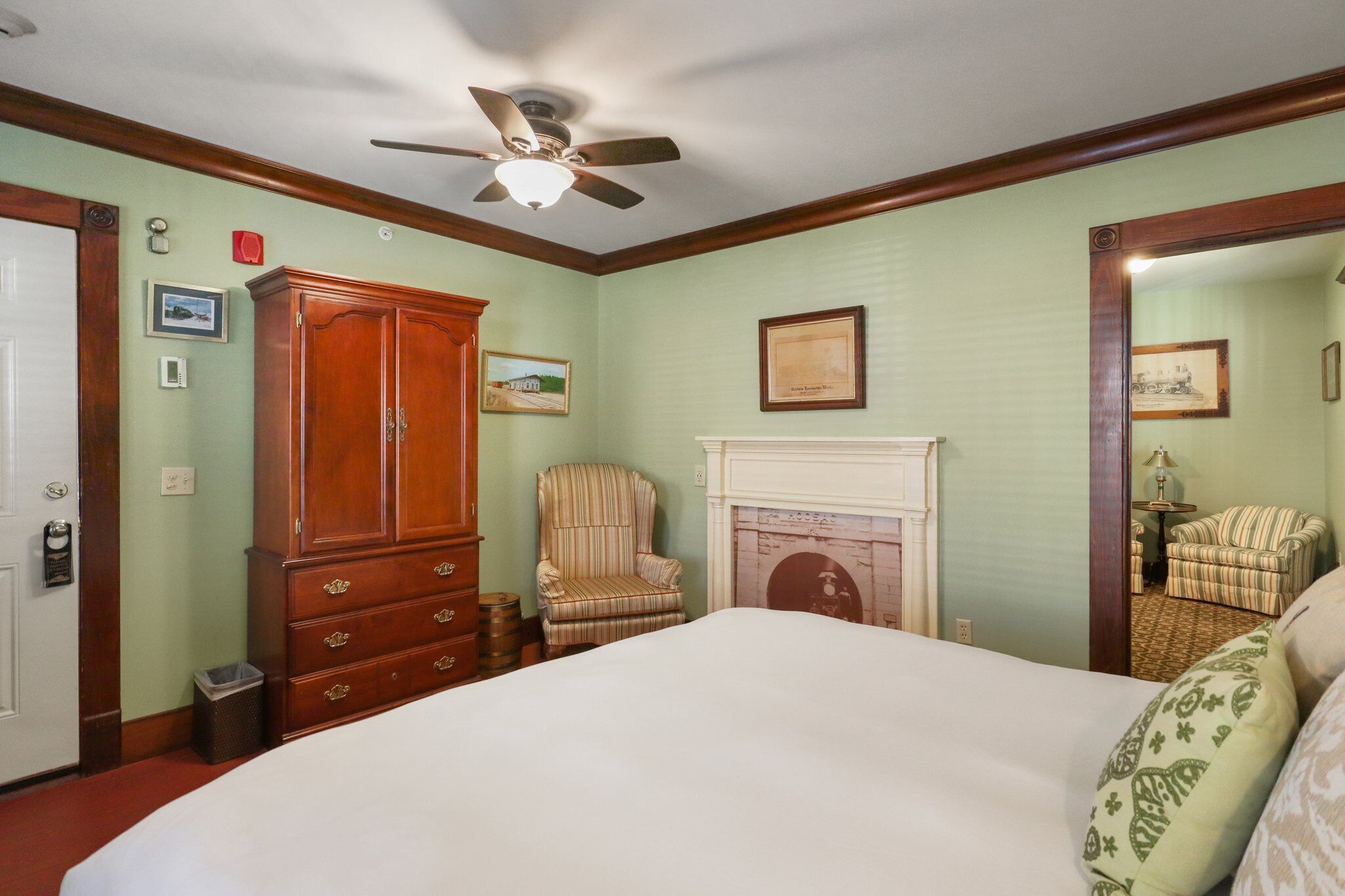 A bedroom with a large bed, a wooden wardrobe, a striped armchair, a fireplace, and a ceiling fan.
