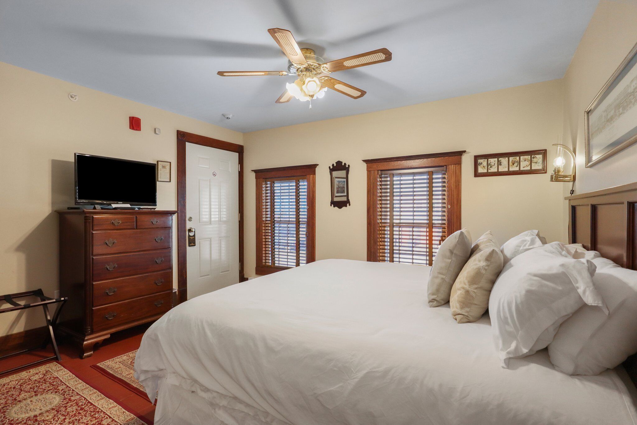 A bedroom with a bed, dresser, television, ceiling fan, and windows.