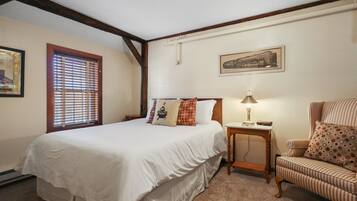 A bedroom with a large bed, a chair, a small table, and a window with blinds.